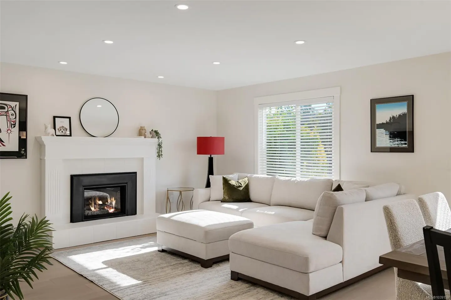 Bright living room with a white sectional sofa, fireplace, red lamp, and large window with blinds.