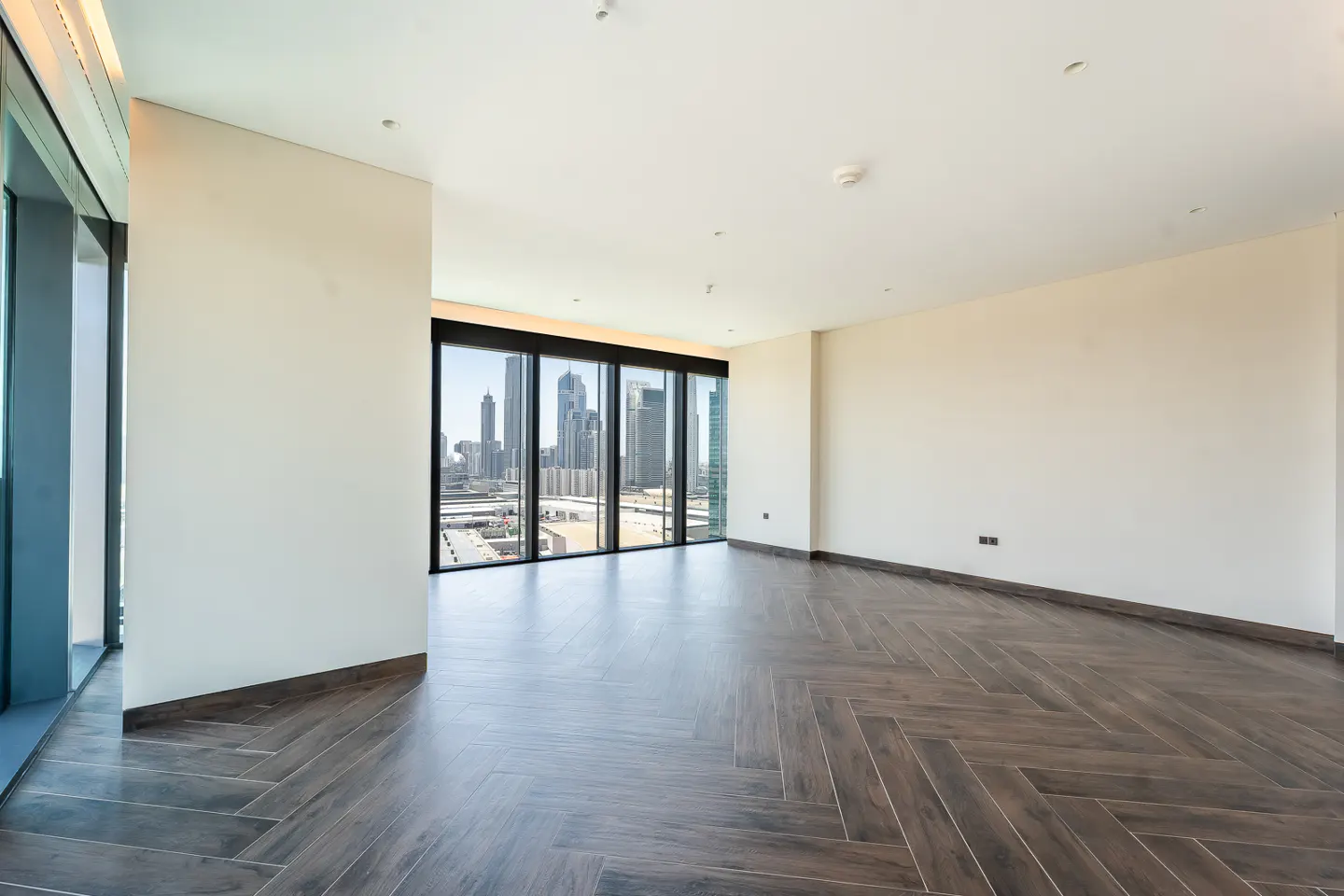 Empty room with herringbone wood floors, white walls, and a large window with a city view.