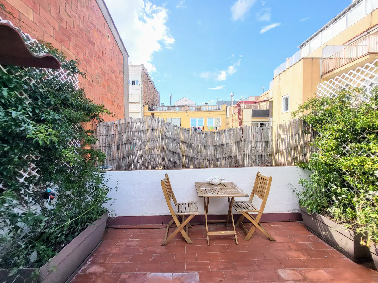 Outdoor patio with red brick tiles, a small wooden table and two chairs, and green plants in planters. A bamboo fence provides privacy.