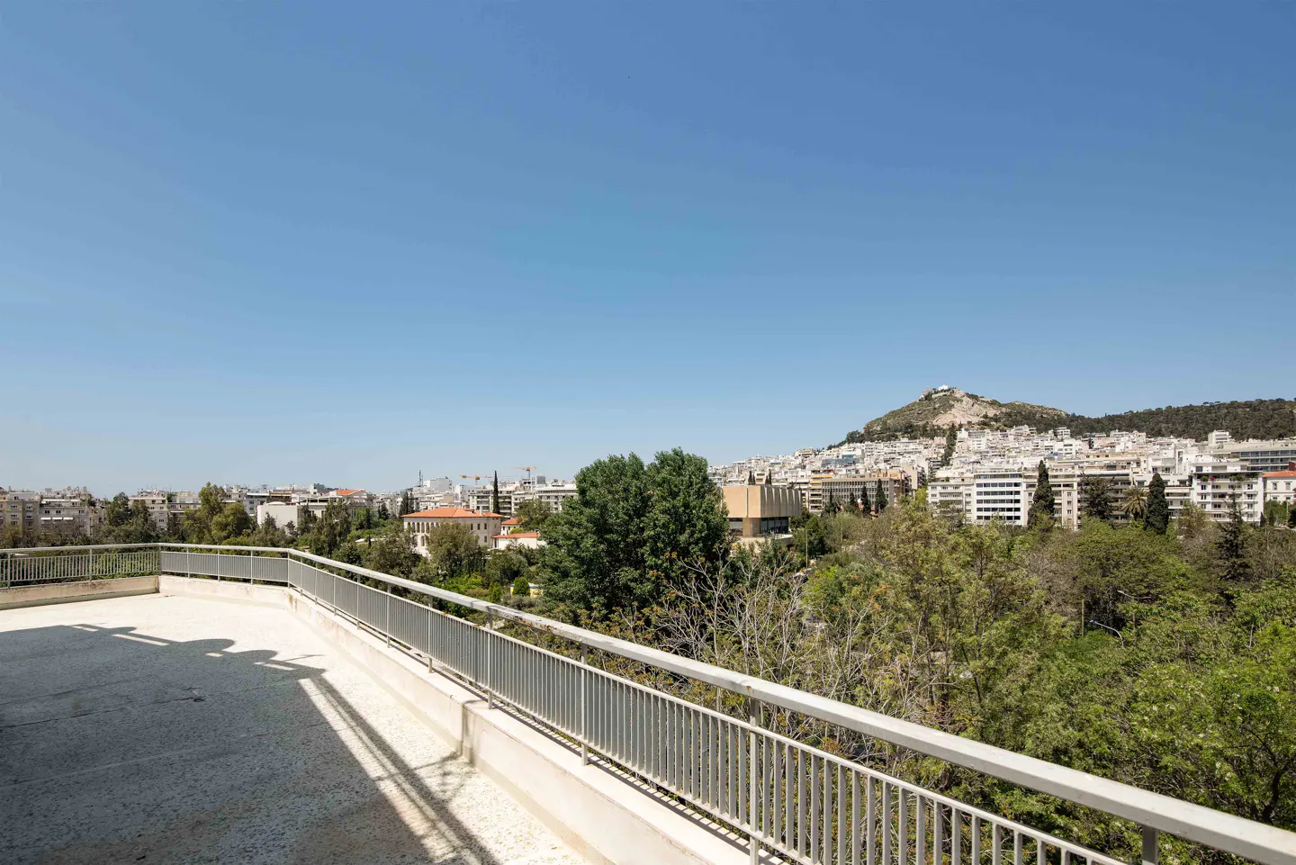 View from a balcony with a gray railing overlooking Athens, Greece, with Mount Lycabettus in the background under a clear blue sky.