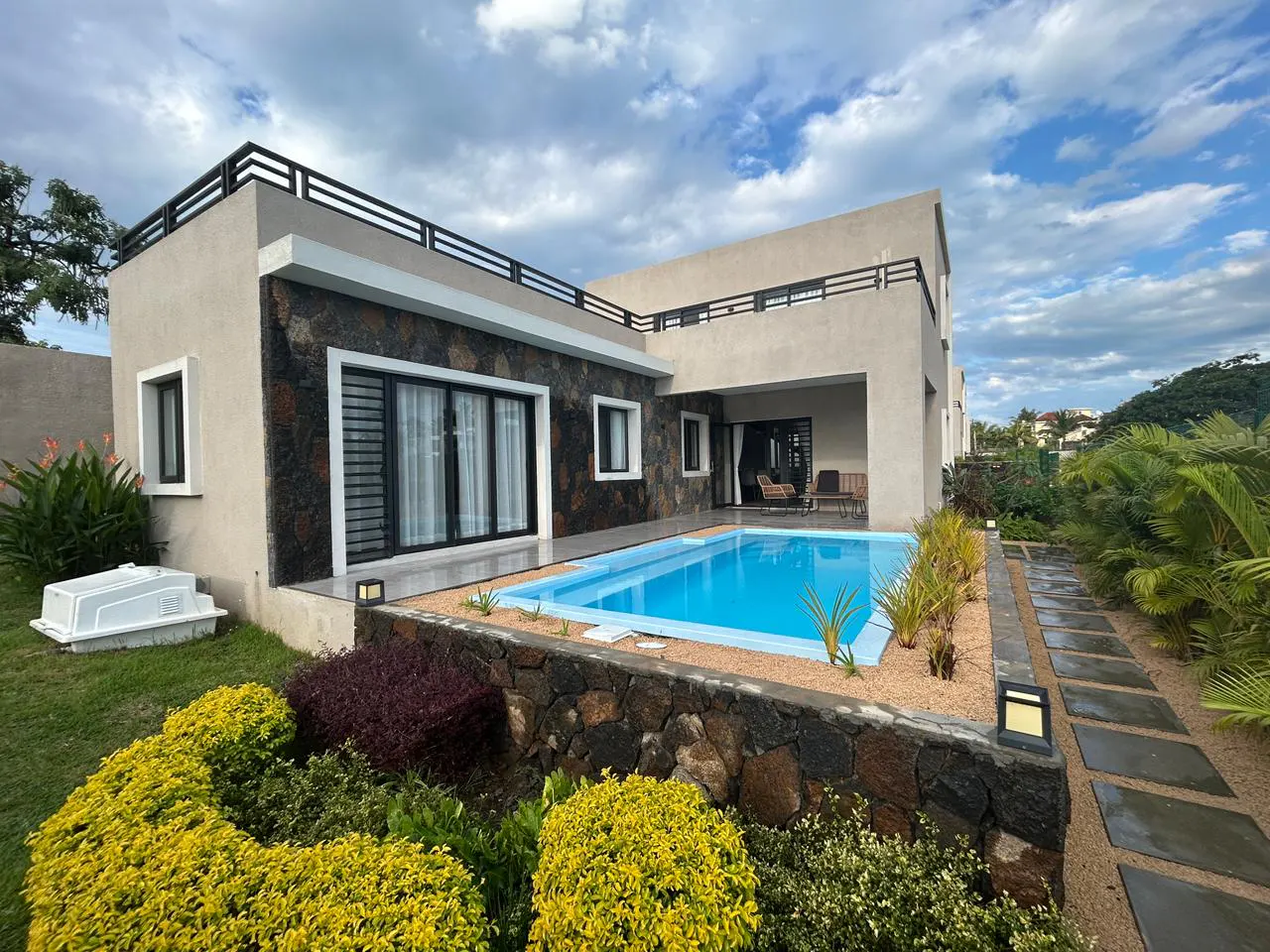 Modern villa with a blue swimming pool, stone accents, and a landscaped garden under a cloudy blue sky.