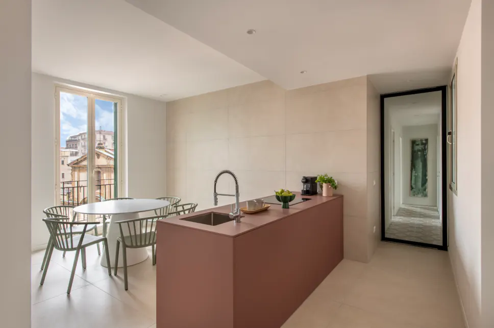 A modern kitchen with a pink island, sink, and black faucet. A white table with green chairs sits near a window with a city view.