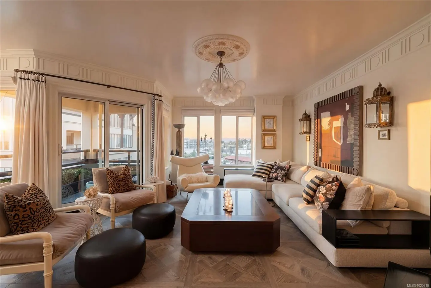 Bright living room with a white sofa, chairs, and a dark wood coffee table. A chandelier hangs from the ceiling. Balcony with sunset view.
