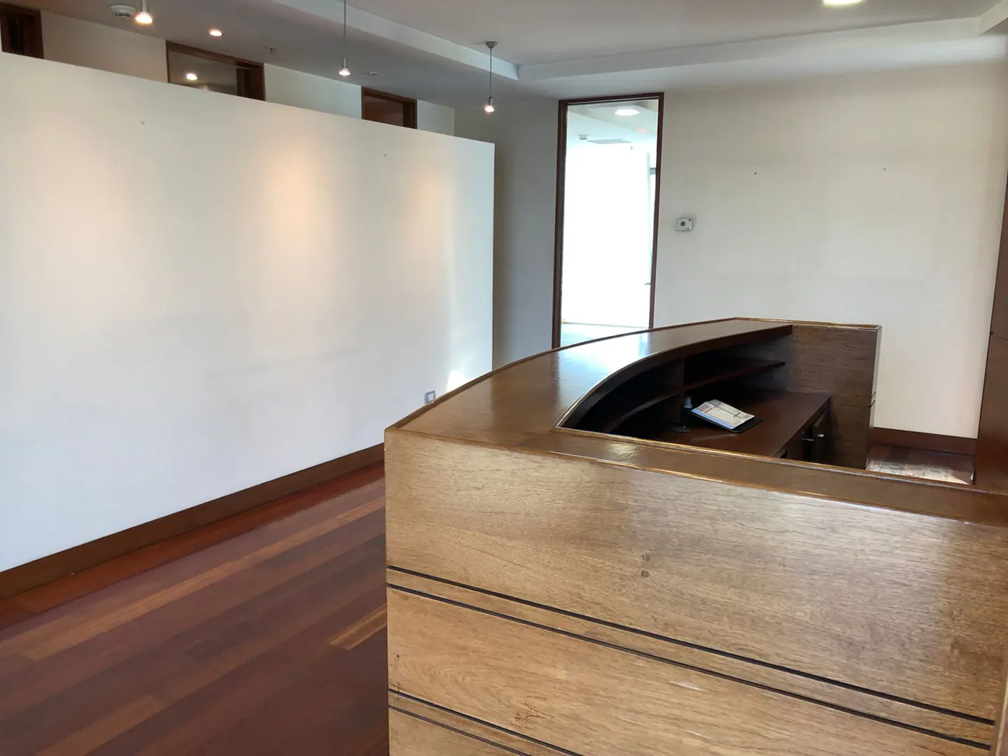 Office space with a wooden reception desk, white walls, and a doorway leading to a bright room. The floor is made of dark wood.