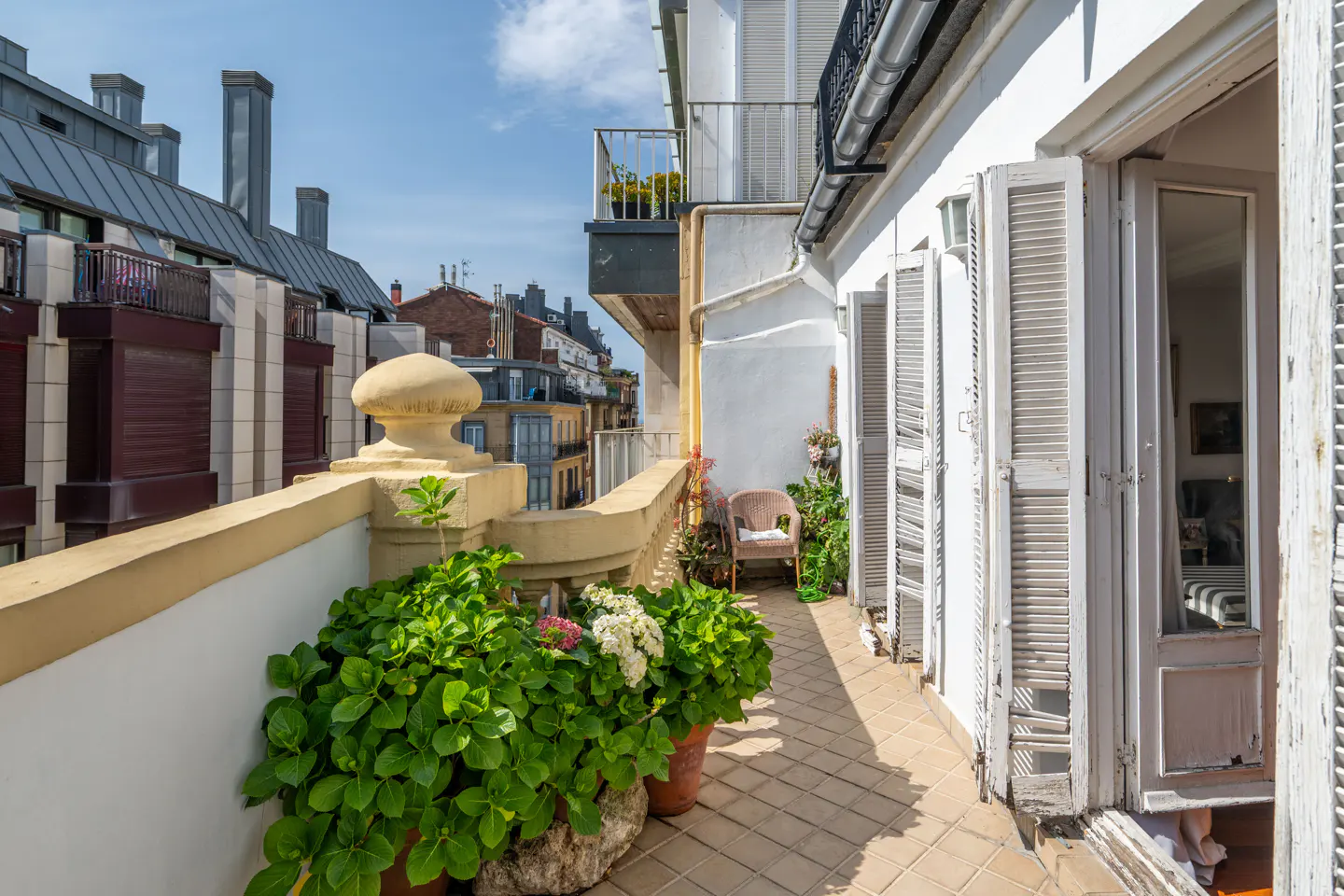 A sunlit balcony with potted plants, a wicker chair, and white shutters. City buildings are visible in the background.