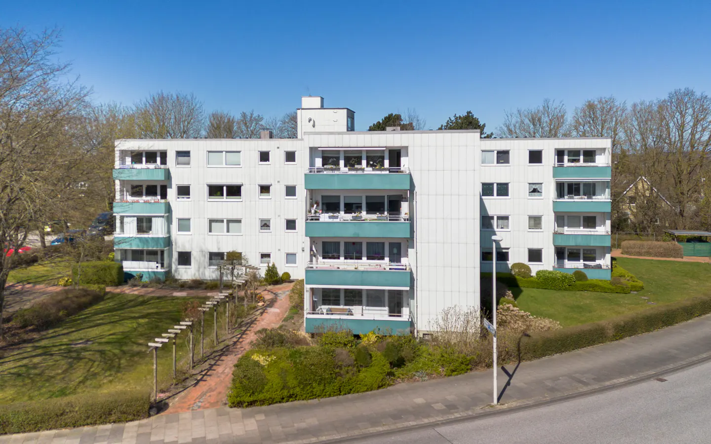 A white apartment building with teal balconies, surrounded by green lawns and trees under a blue sky.