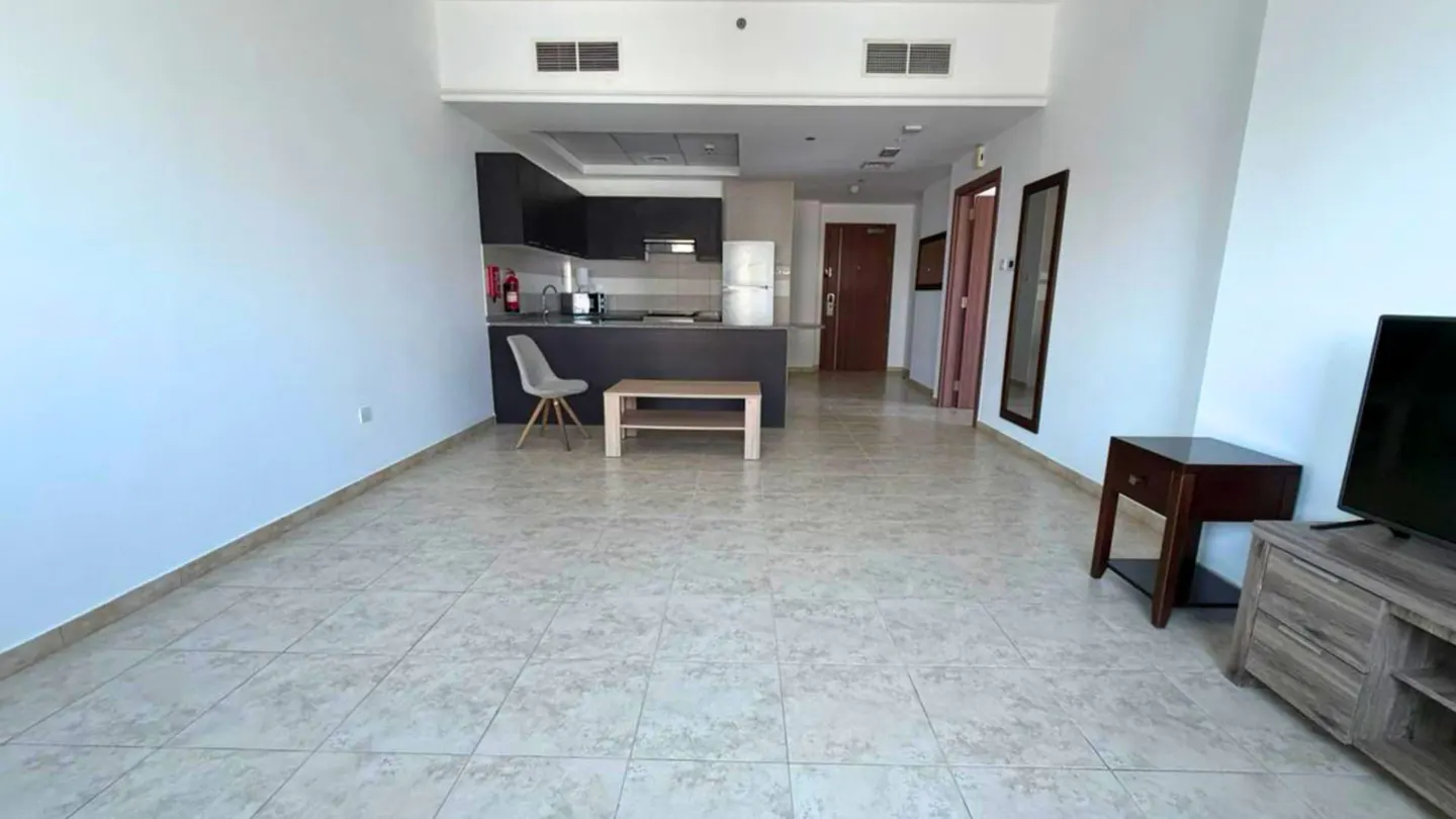 A wide shot of a modern apartment interior with white walls, tile flooring, and an open kitchen with dark cabinets. A light wood coffee table and chair sit in the living area.