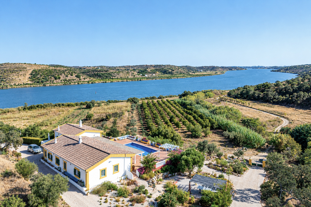 Aerial view of a white villa with a pool, orchard, and gravel driveway overlooking a wide river in a sunny rural landscape. [AI-generated text]