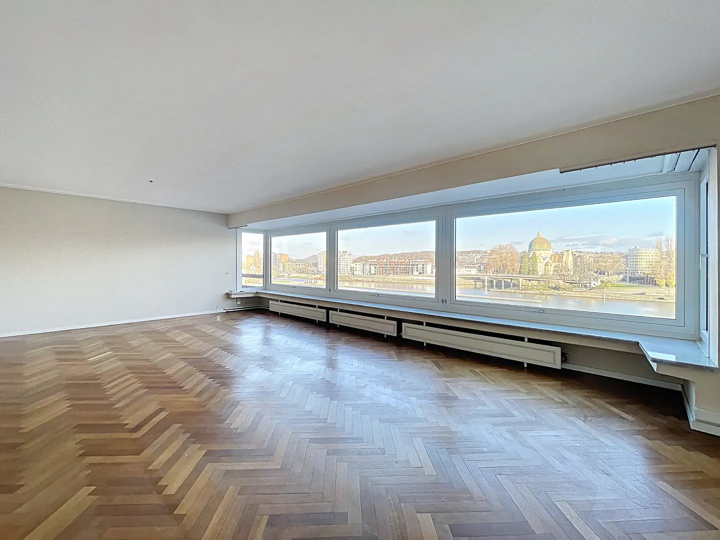 Empty room with herringbone wood floor and large windows overlooking a city and river.
