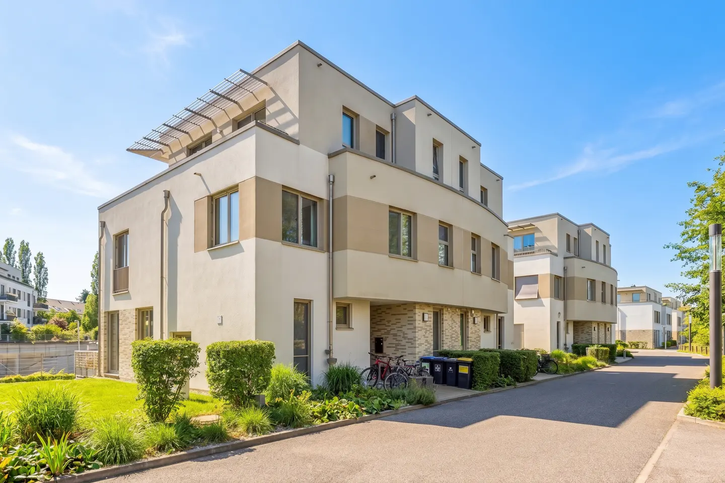 Modern, multi-story townhouses with beige and white exteriors line a paved street under a bright blue sky. Landscaped greenery and bicycles add to the curb appeal.