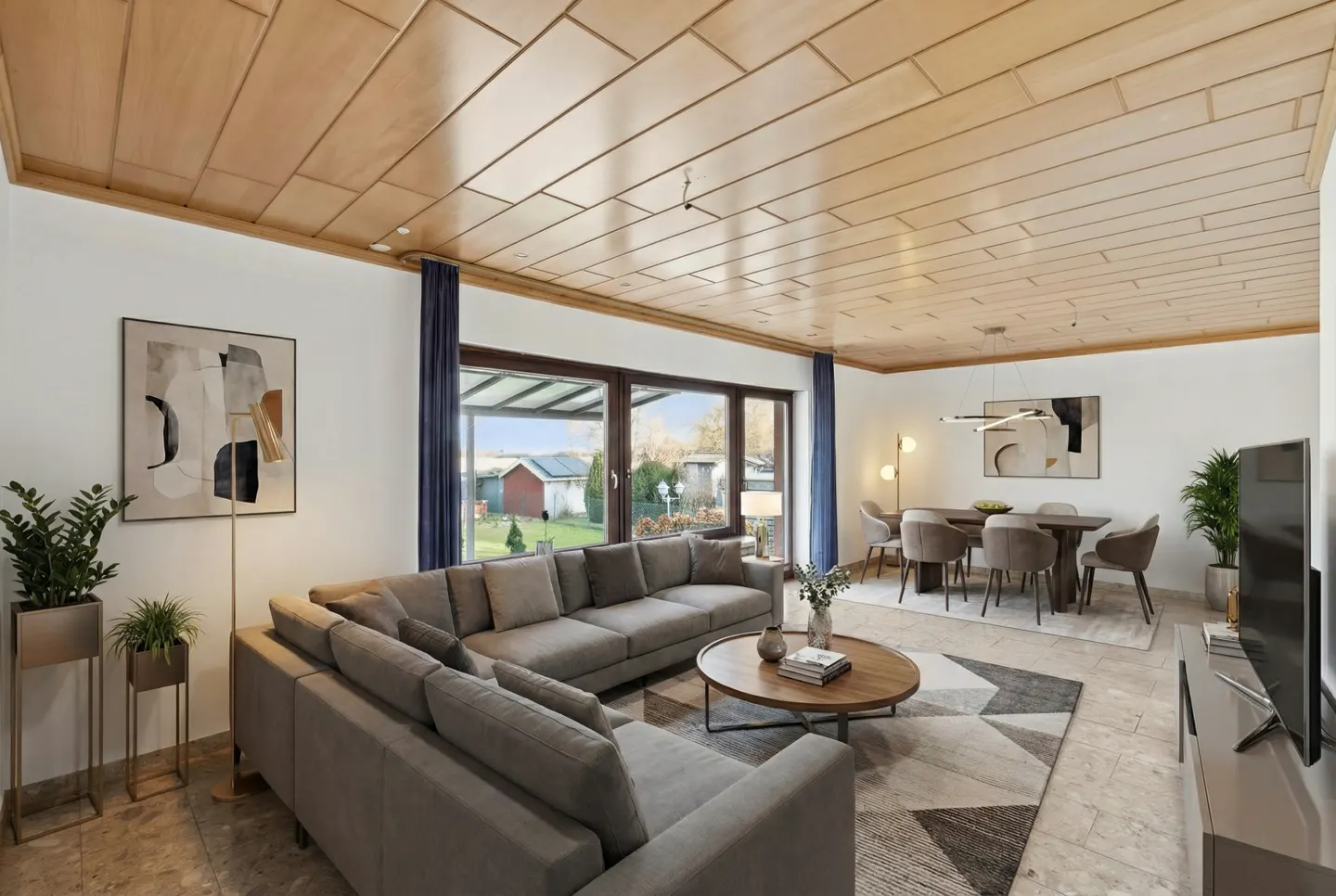 A bright living room with a gray sectional sofa, wood ceiling, round coffee table, and dining area. Large windows offer a view of the outdoors.