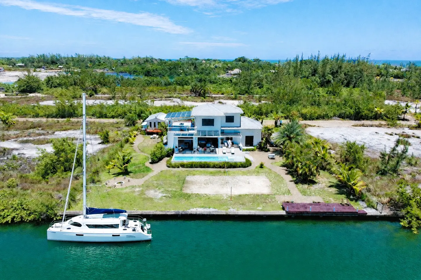 Aerial view of a modern white house with a pool, a sailboat docked on the canal, and lush green trees.