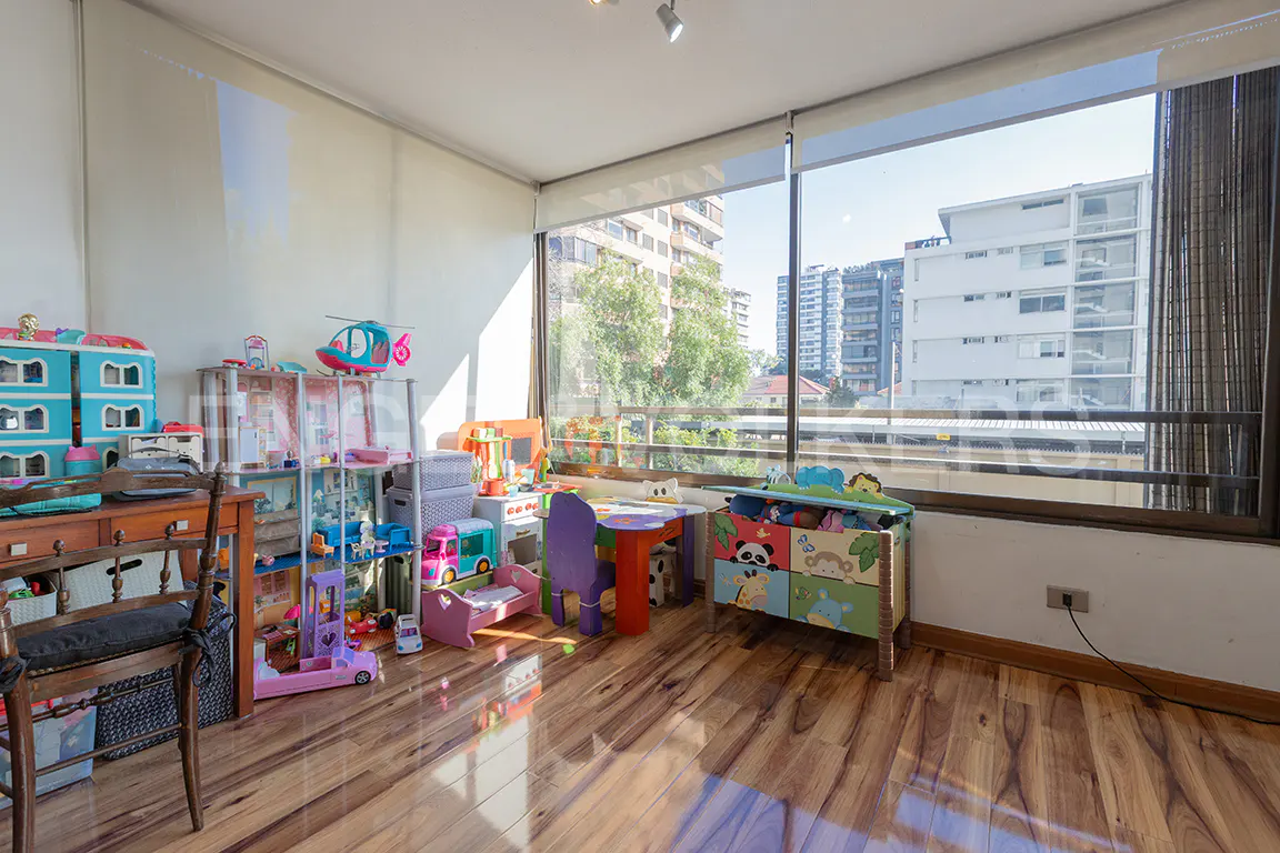 Bright playroom with wood floors, large windows, and city views. Toys, shelves, and a small table fill the space.