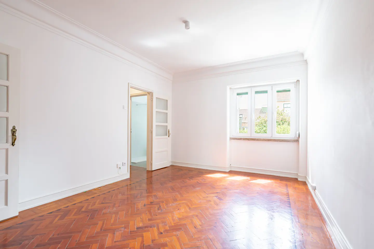 Bright, empty room with white walls, herringbone wood floor, and a window. A doorway leads to another room.
