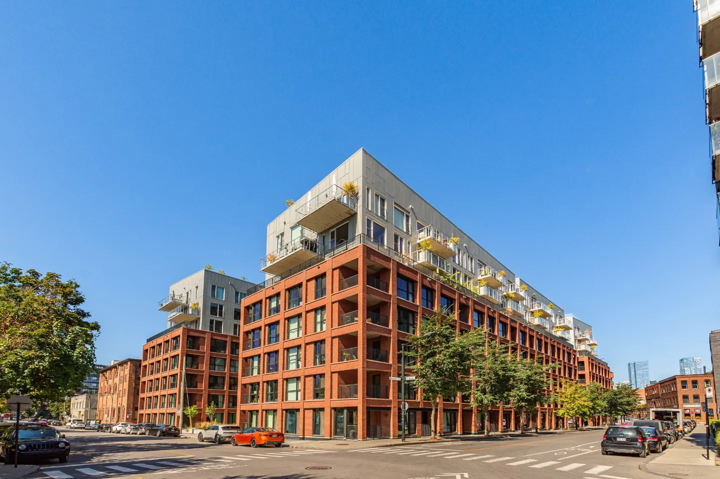 Modern brick apartment buildings with balconies line a city street under a clear blue sky. Cars are parked along the street.
