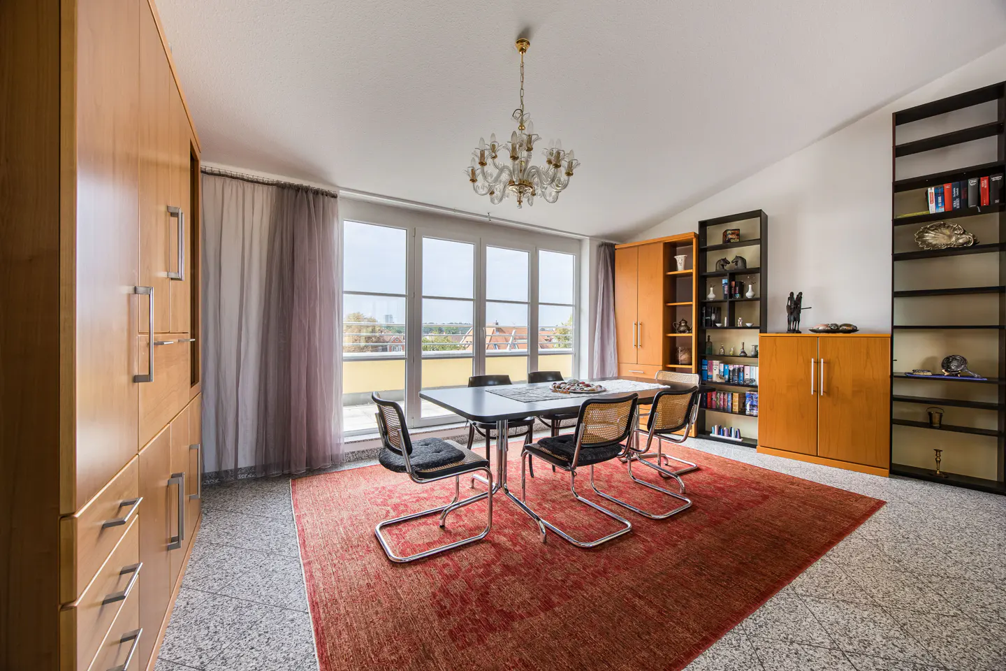 Bright dining room with a table, six chairs, and a red rug. Large windows offer a city view. Built-in cabinets and bookshelves line the walls.