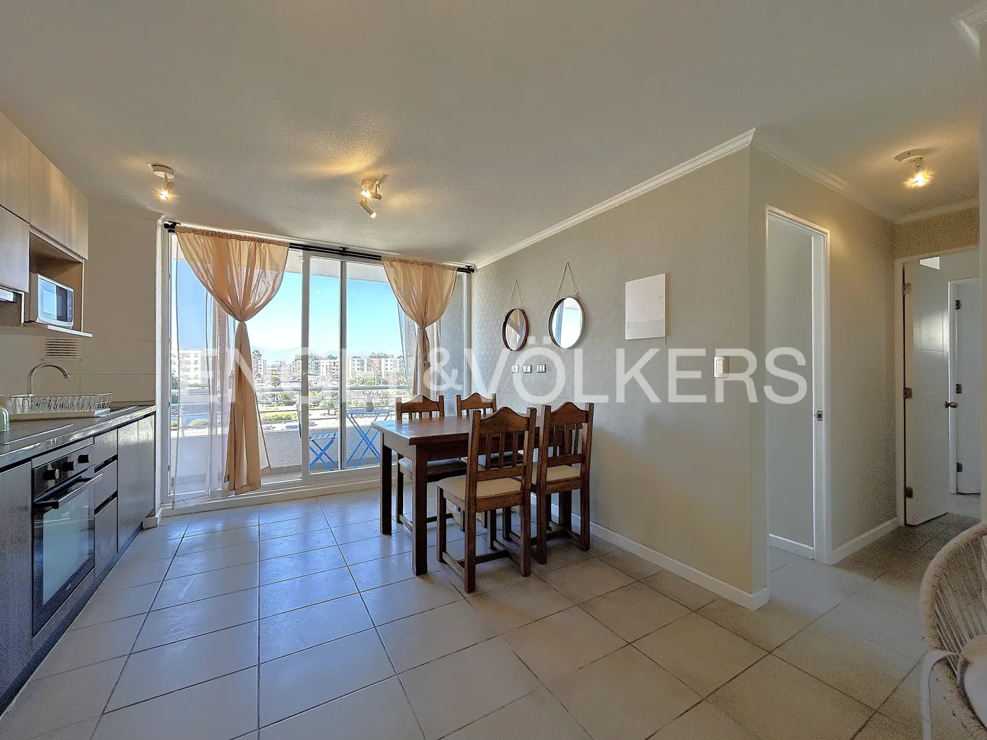 Bright apartment interior with kitchen, dining table, and balcony overlooking a cityscape. Neutral tones and tile flooring.