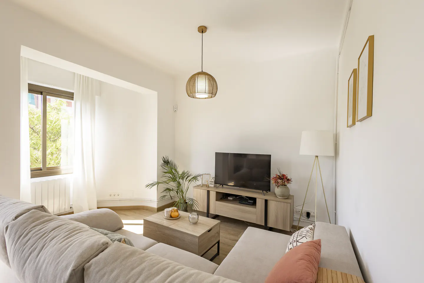 Bright living room with beige sectional sofa, wood coffee table, TV stand, and a window with sheer white curtains.