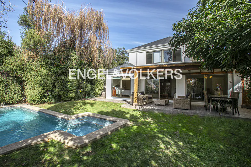 Backyard of a white two-story house with a swimming pool, patio furniture, and lush green trees under a blue sky.