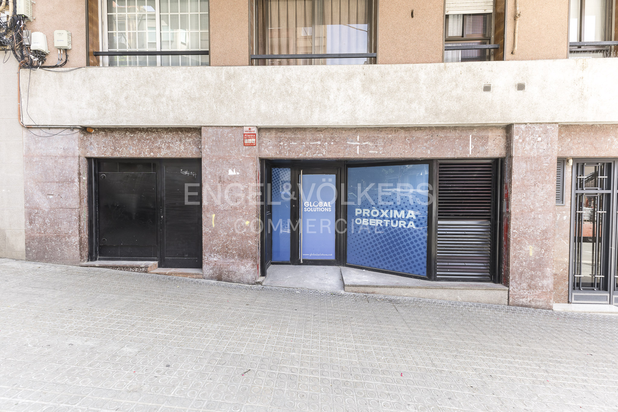 Commercial storefront with a blue "Pròxima Obertura" sign on a stone-clad building facade along a paved street.