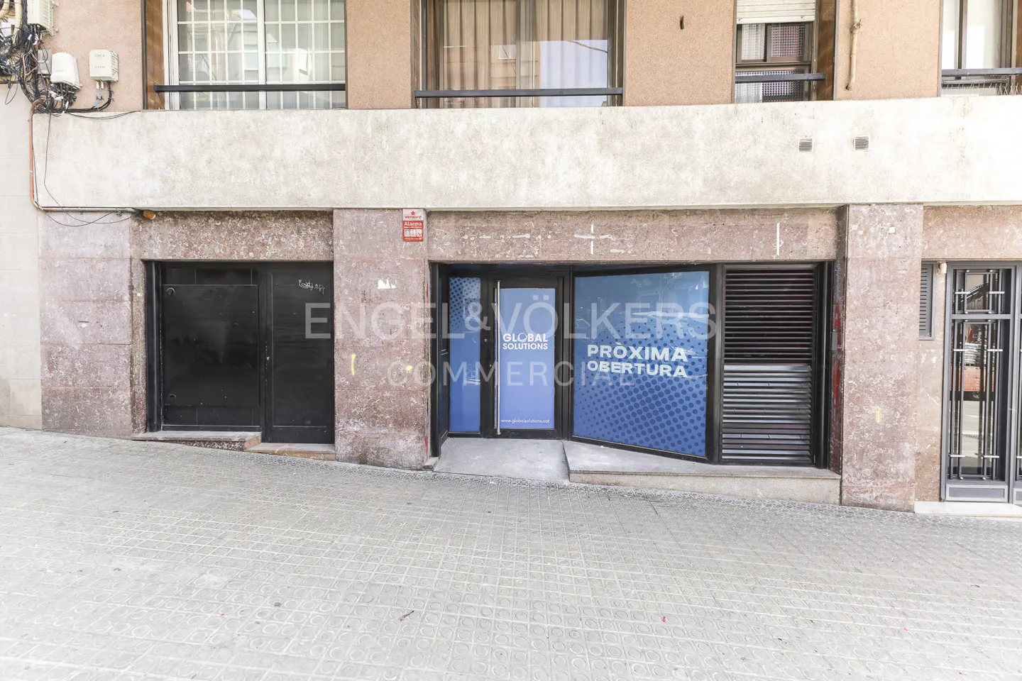 Commercial storefront with a blue "Pròxima Obertura" sign on a stone-clad building facade along a paved street.