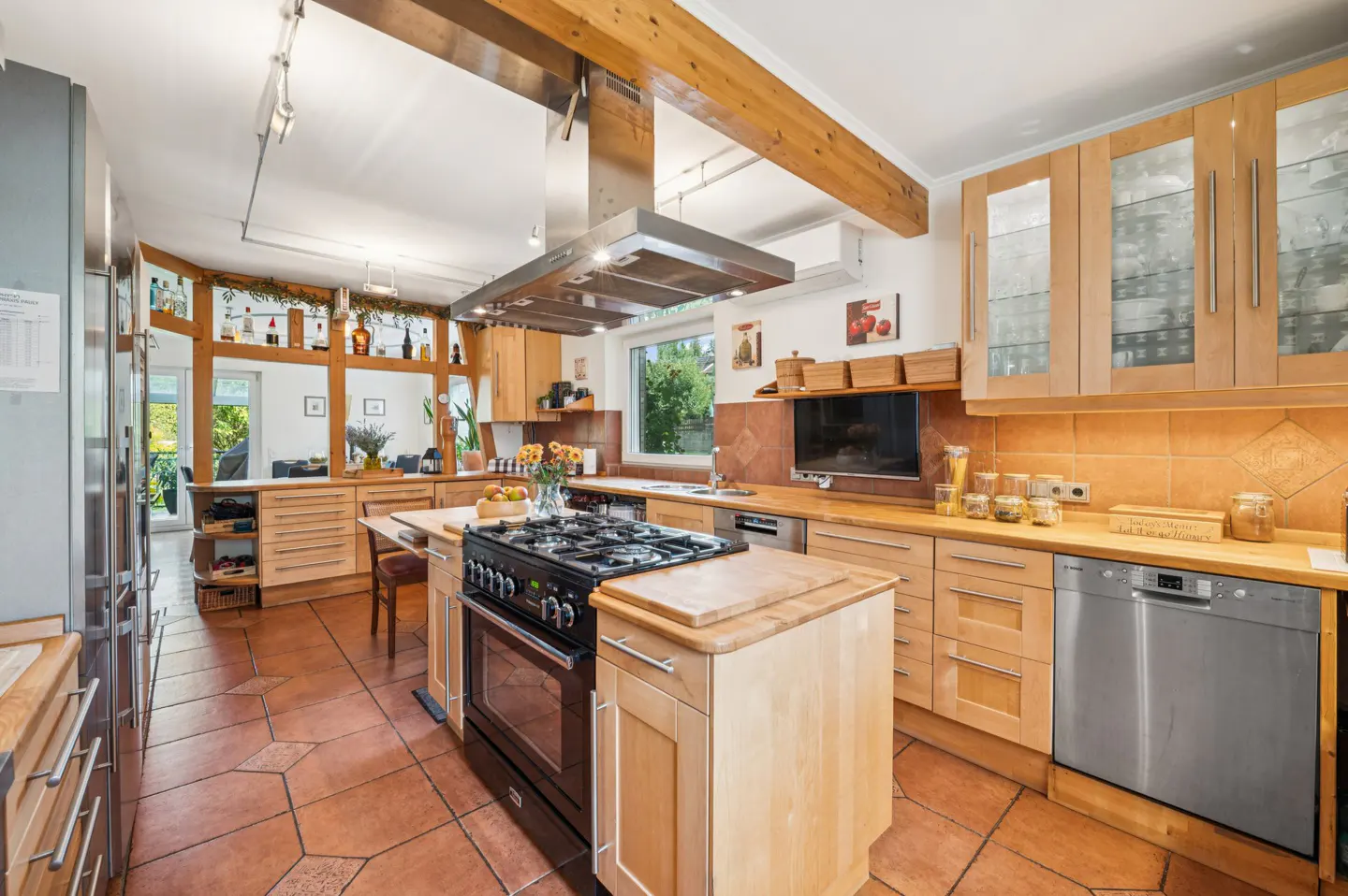 A bright kitchen with light wood cabinets, a black stove on an island, and terracotta tile floors. A stainless steel range hood hangs above the stove.