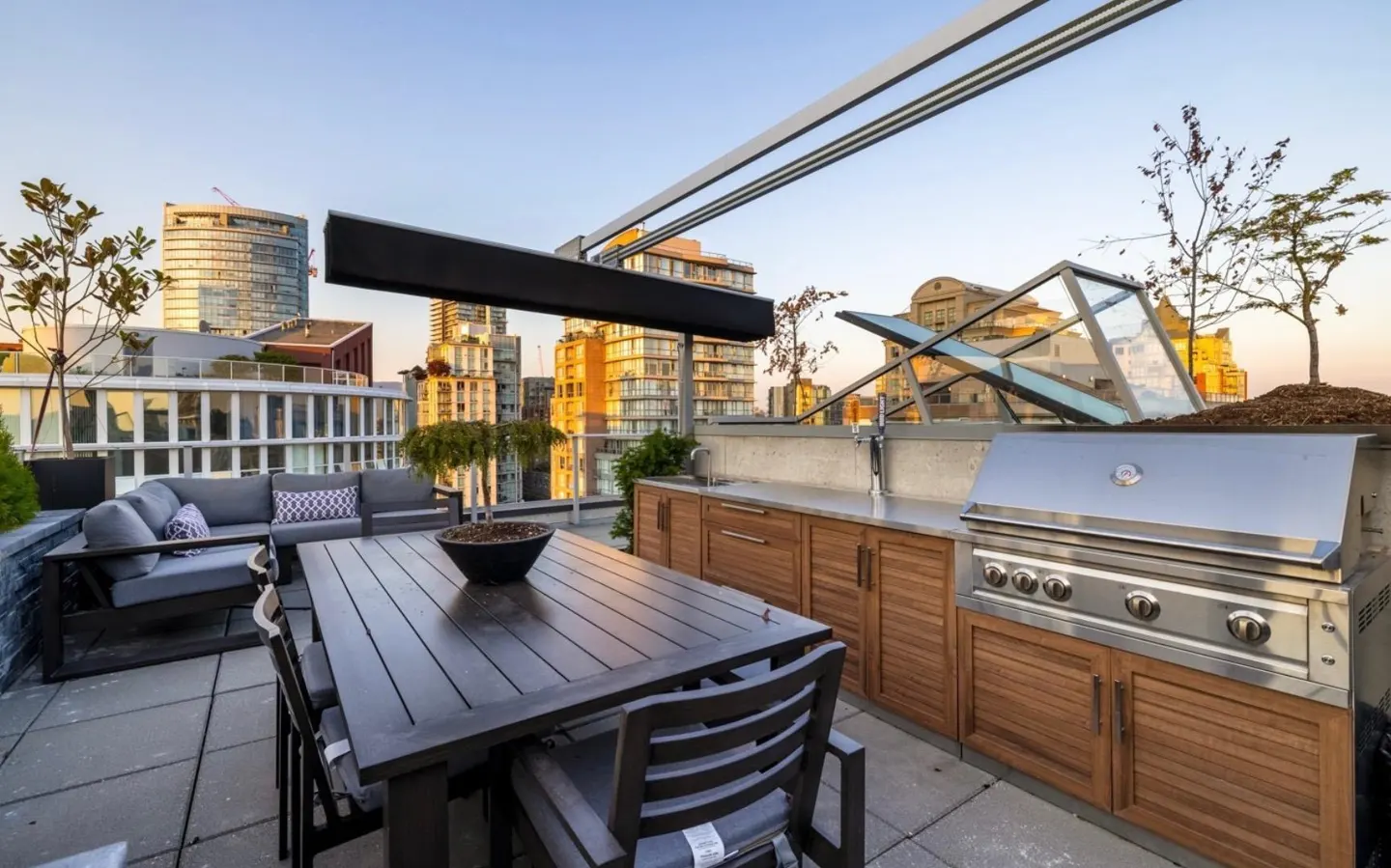 Rooftop patio with a grill, dining table, and sofa. City buildings are visible in the background.