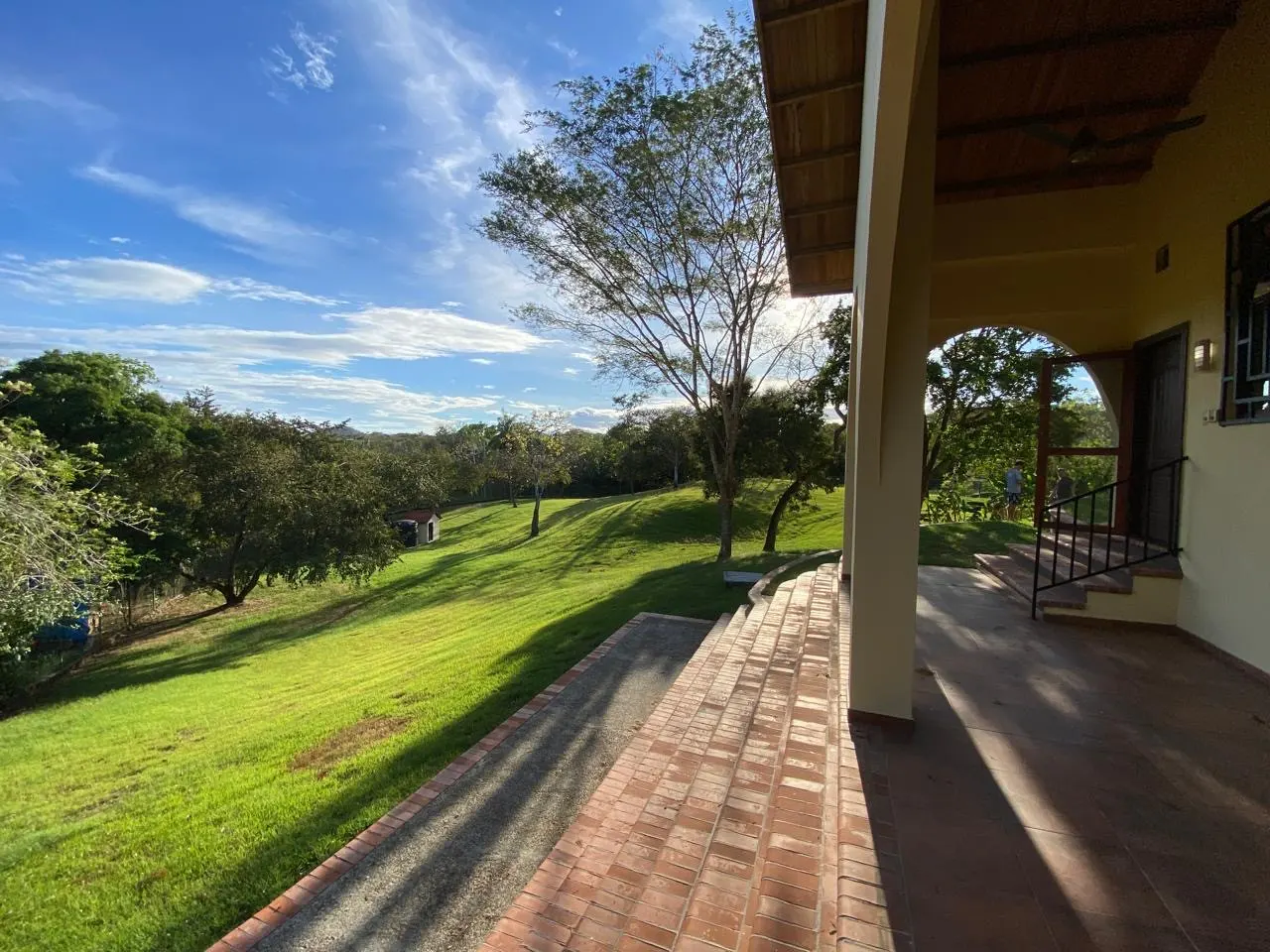 View from a porch with brick steps and a green lawn with trees under a blue sky.