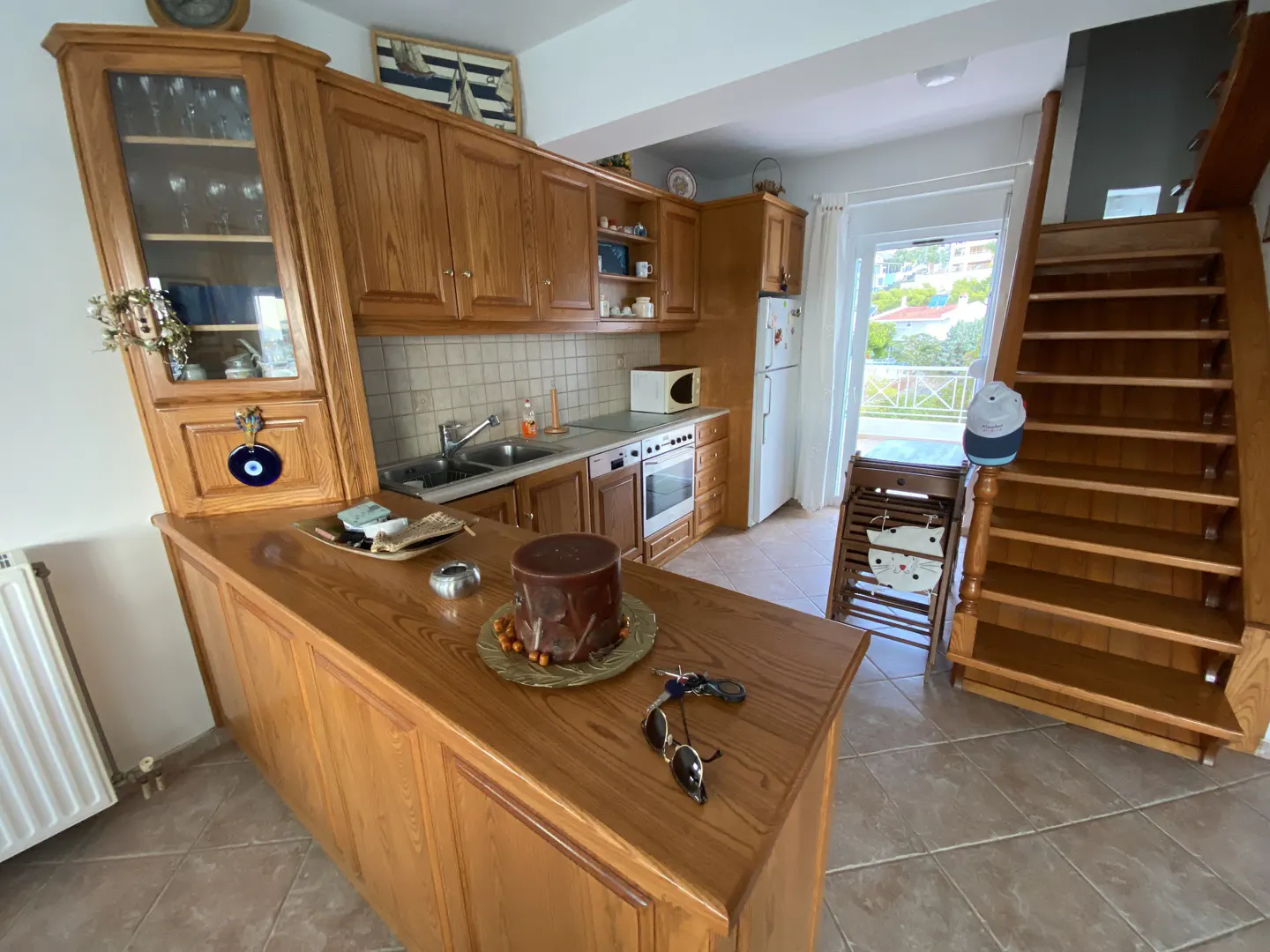 A sunlit kitchen with wood cabinets, a breakfast bar, and stairs. A white fridge and oven are visible. Sunglasses and keys rest on the counter.