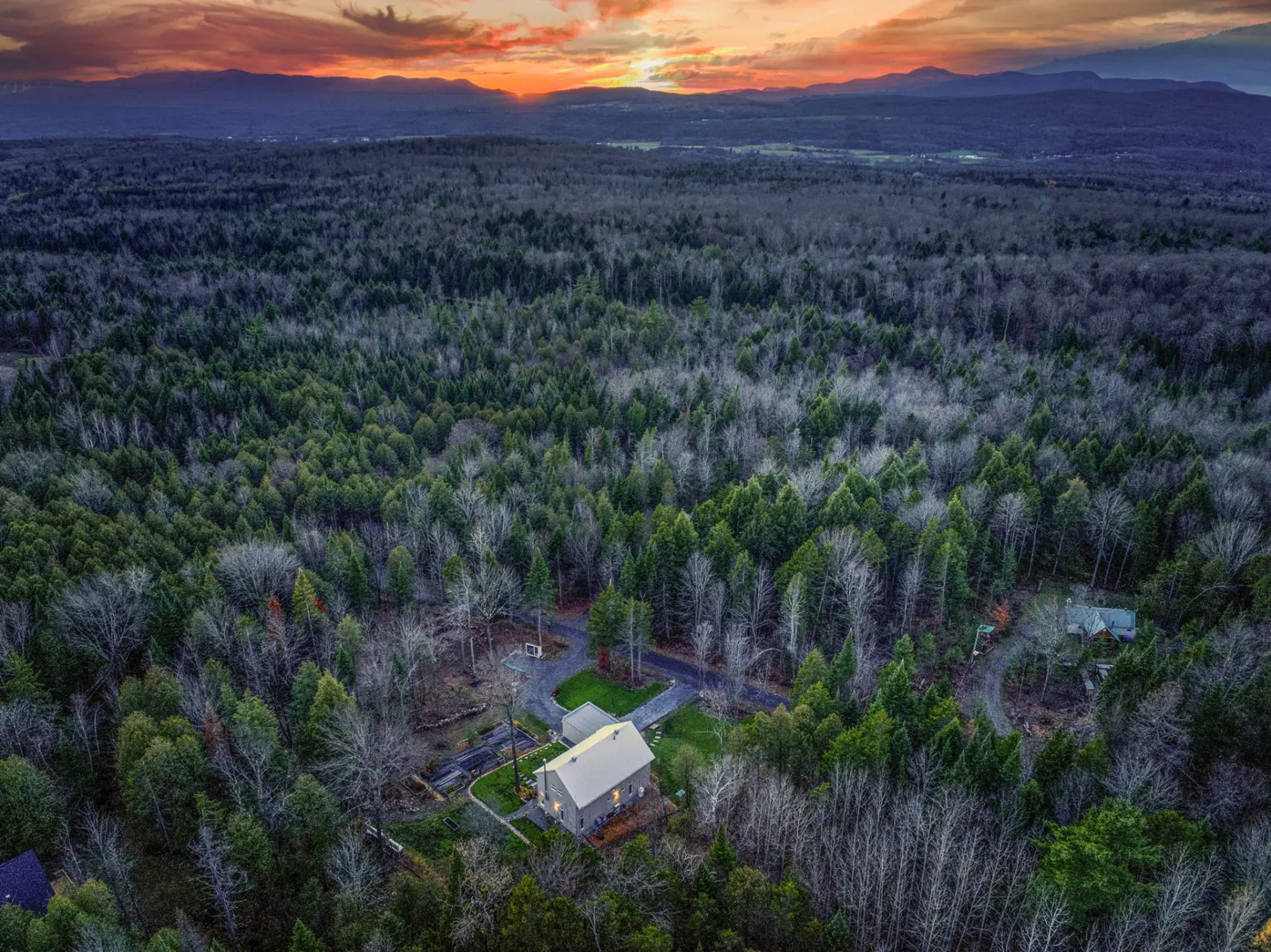 Aerial view of a gray house with a silver roof surrounded by a dense forest at sunset.