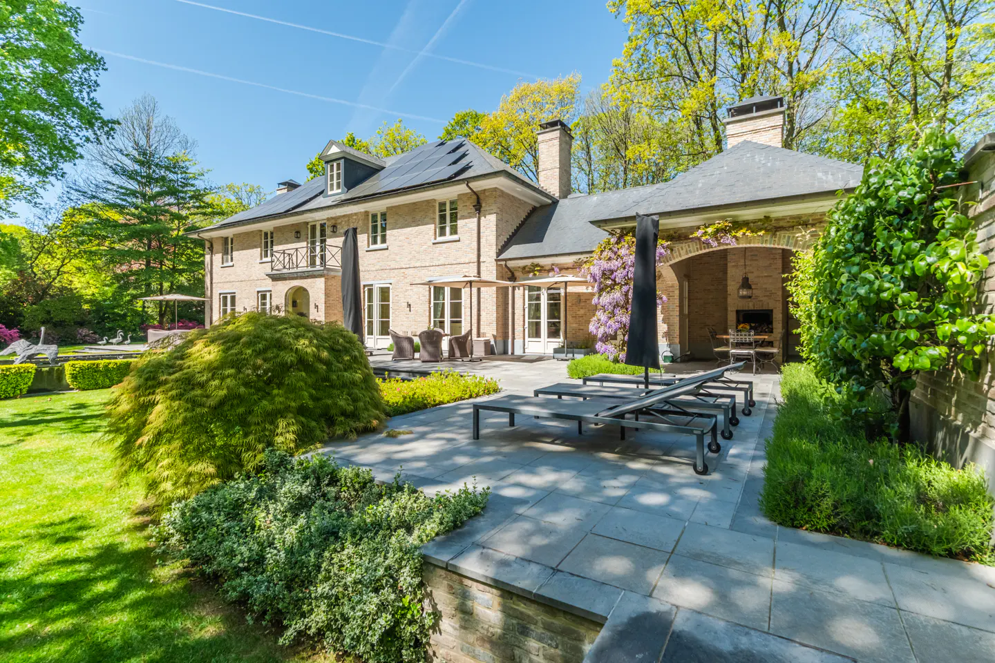 Backyard view of a large tan brick house with a stone patio, lounge chairs, and green landscaping on a sunny day.