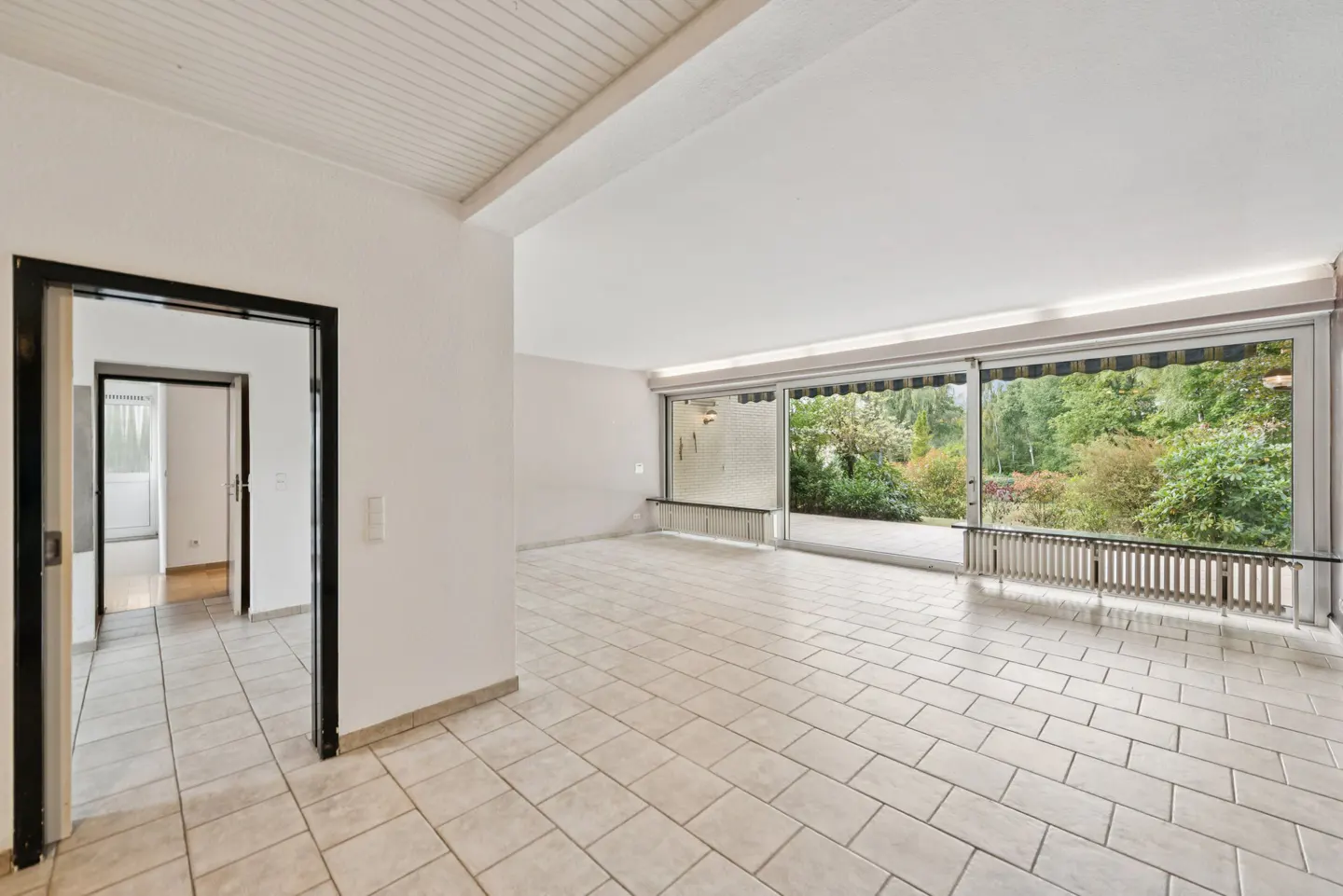 Bright, empty room with white walls, tiled floor, and black-framed doorway. Large sliding glass doors open to a balcony with a view of lush greenery.