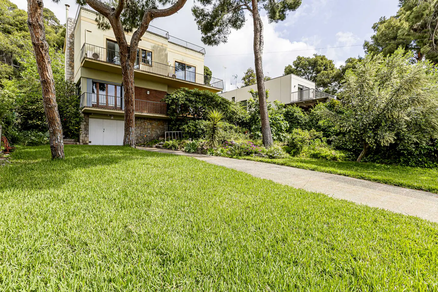 Two-story house with balconies, a stone foundation, and a white garage door, surrounded by green lawn and trees.