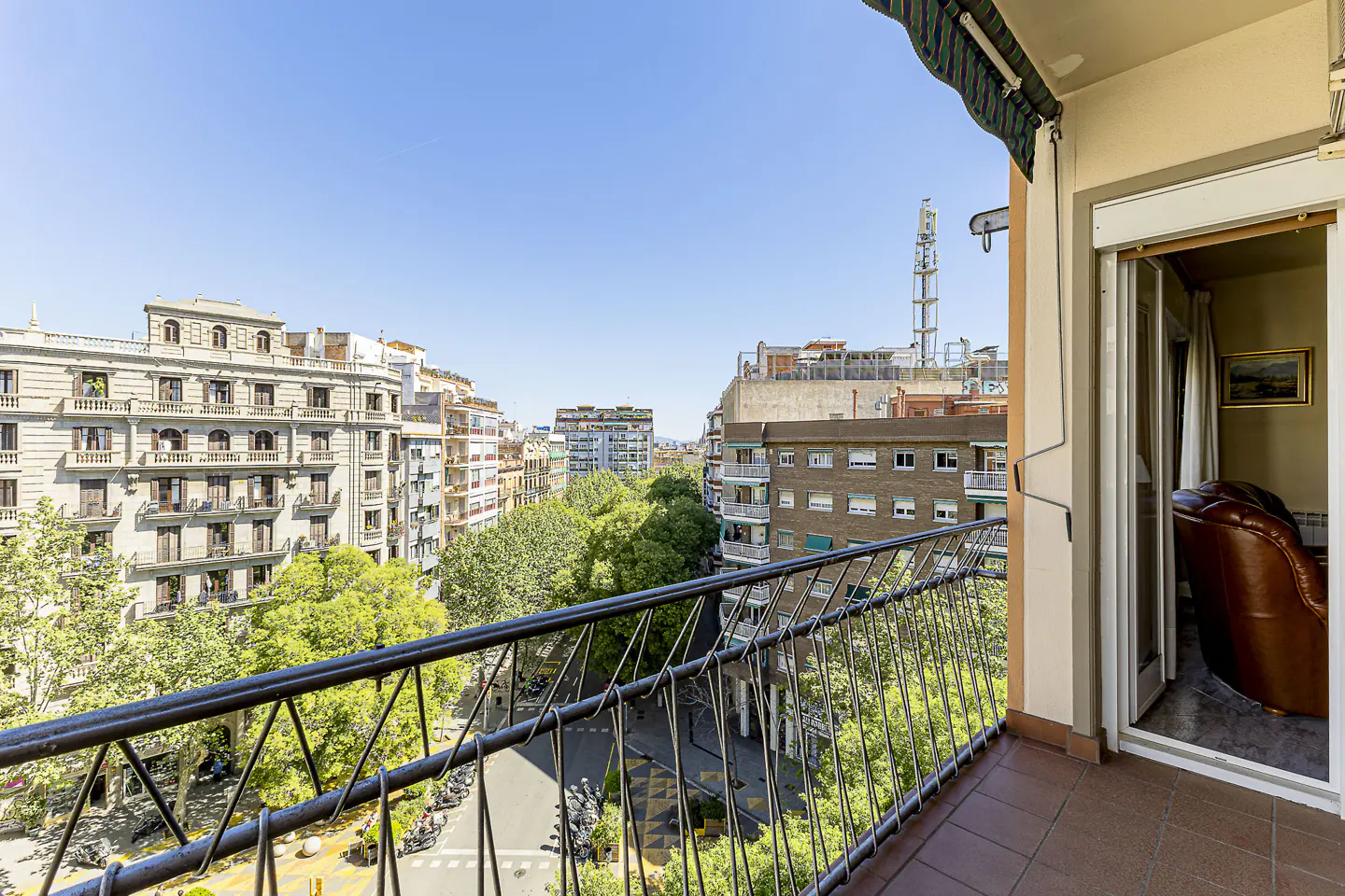 View from a balcony with black railings overlooking a city street with trees and buildings under a blue sky. A glimpse of a brown leather chair is visible through an open door.