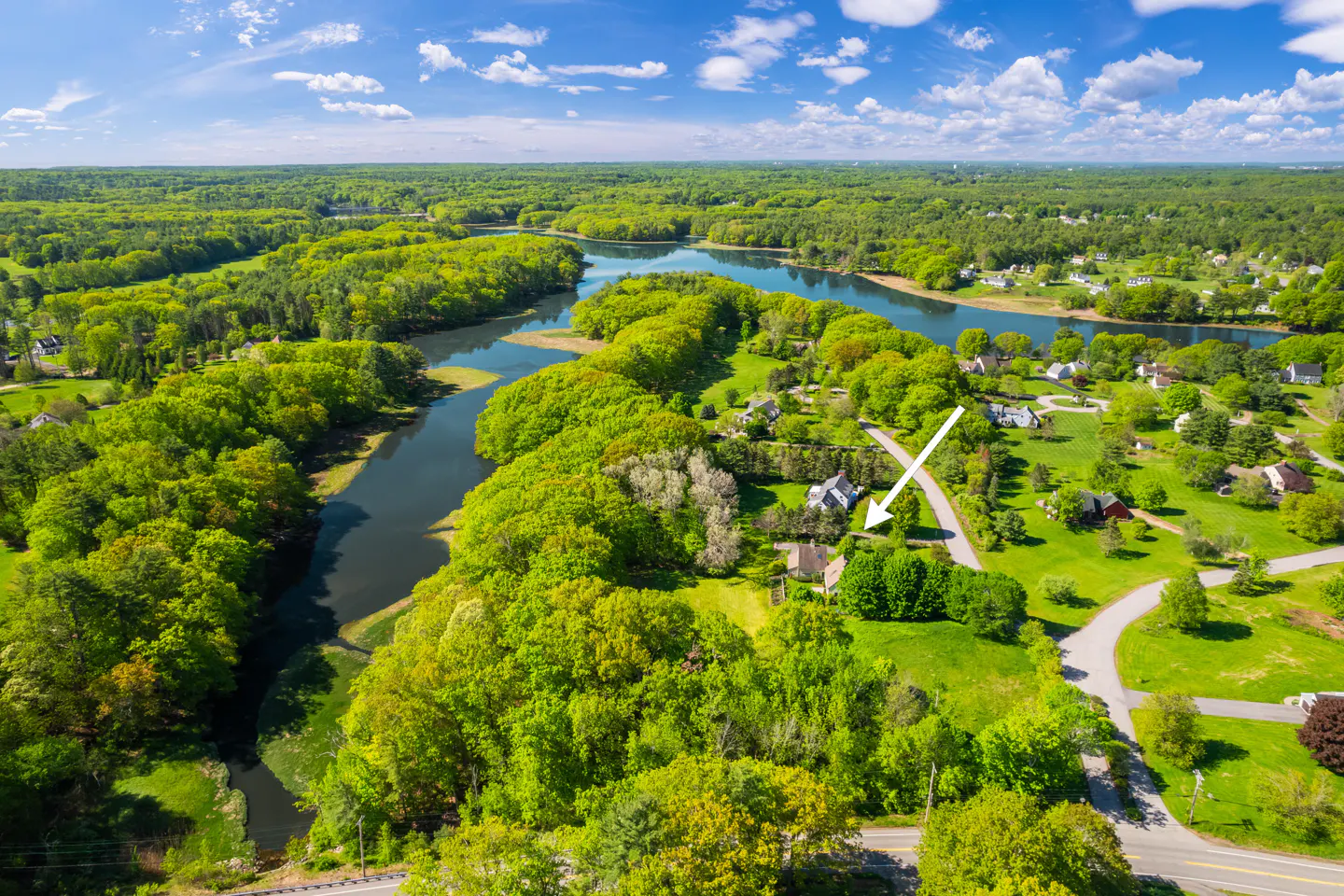 Aerial view of a house nestled in a lush, green landscape next to a winding river under a bright blue sky.