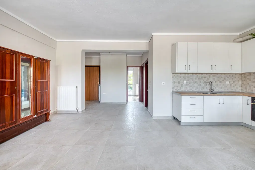 Bright, empty apartment with gray tile floors, white walls, and white kitchen cabinets. A large, dark wood wardrobe stands against the left wall.