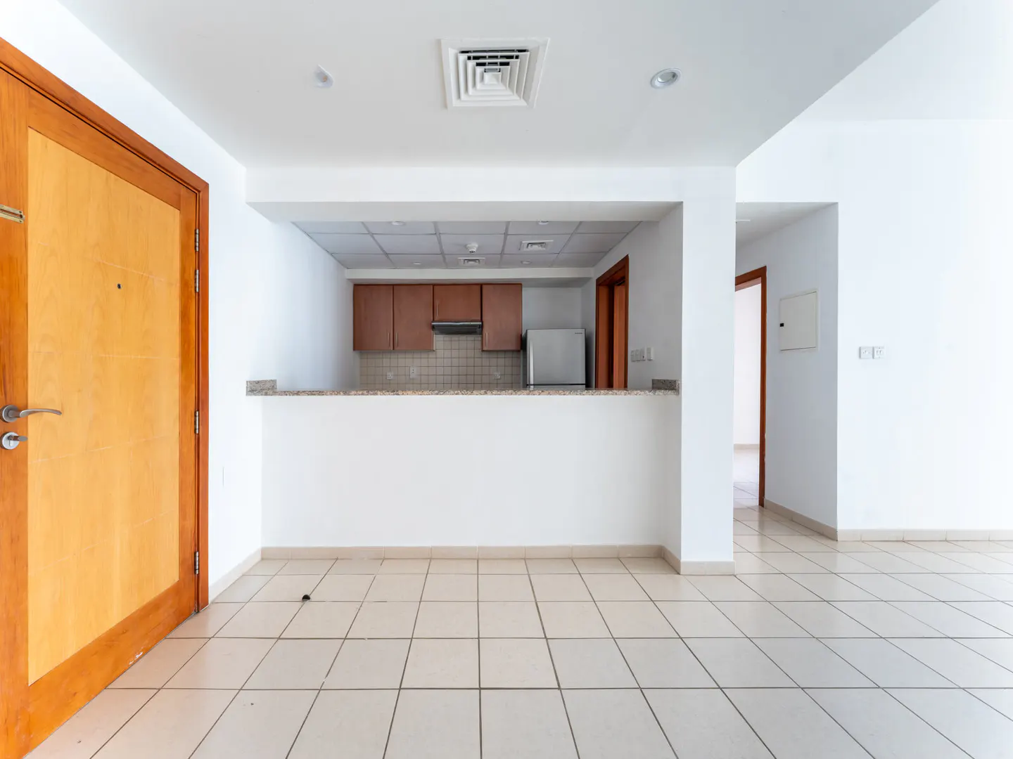 An empty room with white walls and beige tile floors. A wooden door is on the left, and a kitchen with brown cabinets is in the background.