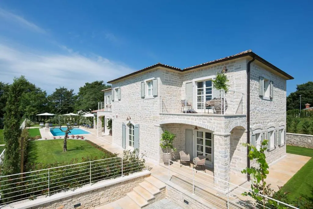 Two-story stone house with a pool and green lawn under a blue sky. Balconies with chairs and tables. White fence and steps.