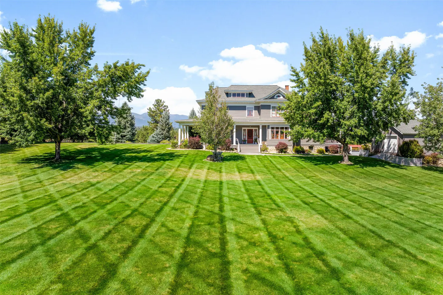 Two-story gray house with a large green lawn and trees under a blue sky.