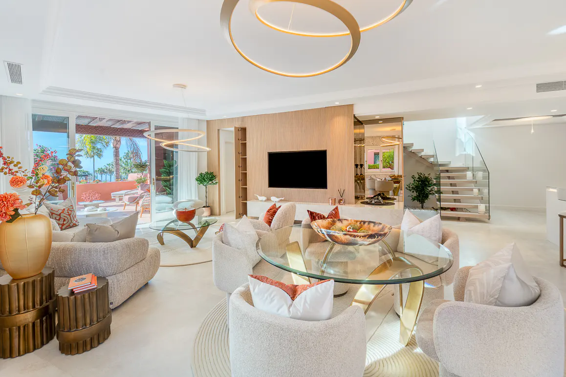 Bright, modern living room with white walls, beige furniture, and gold accents. A glass table sits in the center, with a view of a patio and ocean.