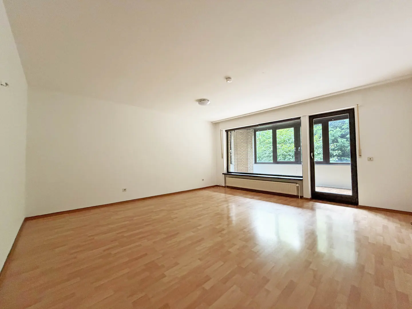Empty living room with light wood flooring, white walls, large windows, and a glass door leading to a balcony.