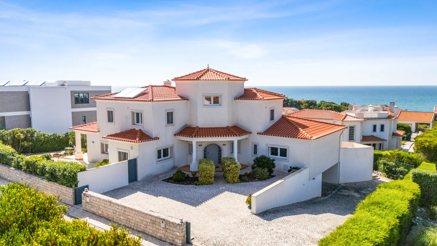 Aerial view of a white, multi-story house with a red tile roof near the ocean under a blue sky.