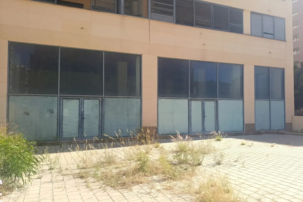 Exterior view of a vacant commercial space with large windows and overgrown weeds on a paved area.