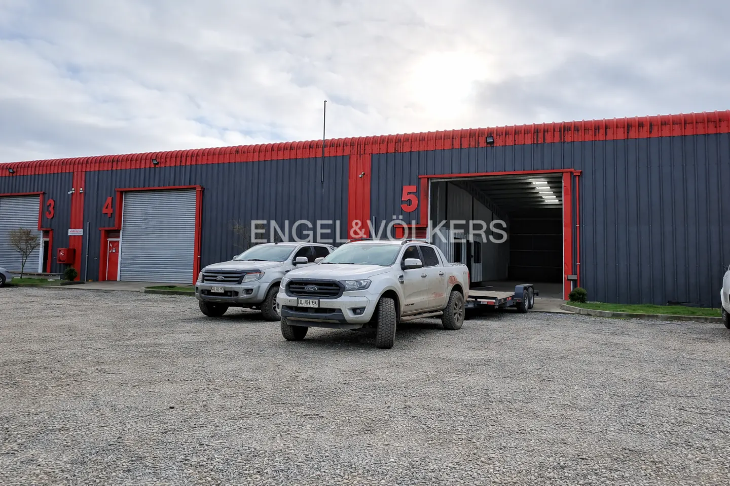 Two white pickup trucks parked in front of a grey and red industrial warehouse with numbered bays on a gravel lot.
