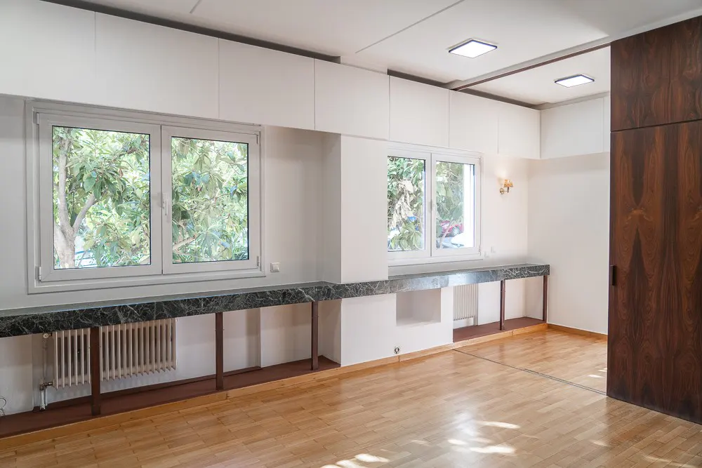 Bright room with wood floors, white walls, and two windows showing green trees. A dark marble counter runs below the windows.