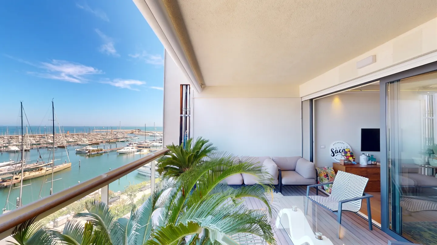 Modern balcony with lounge seating and potted plants overlooking a marina filled with sailboats under a clear blue sky.