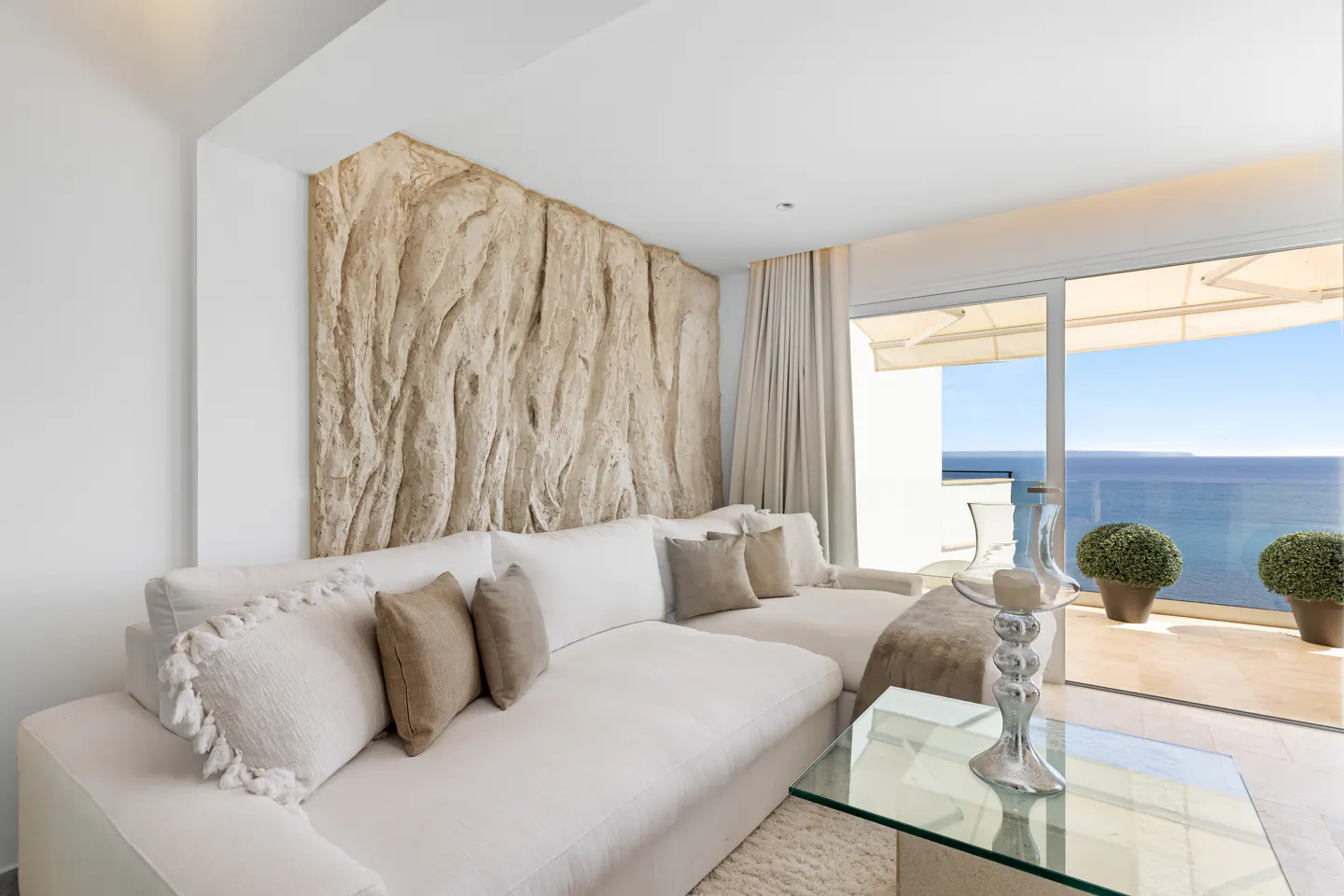 Bright living room with white sectional sofa, textured wall art, and ocean view through sliding glass doors.
