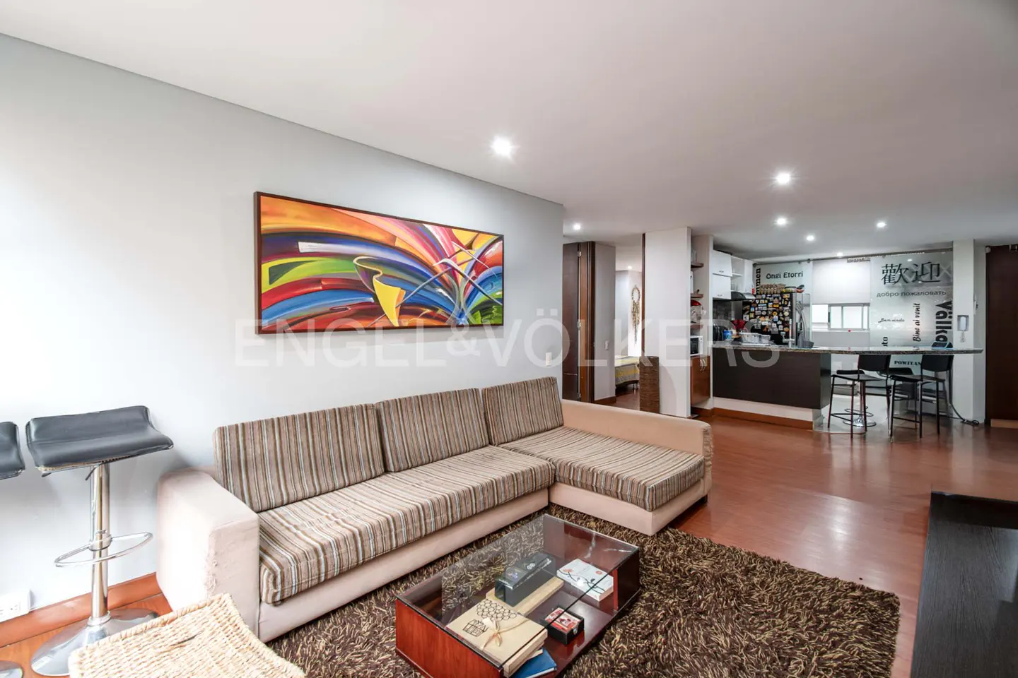 Living room with a striped sofa, glass coffee table, and colorful abstract art. Kitchen bar with stools in the background.