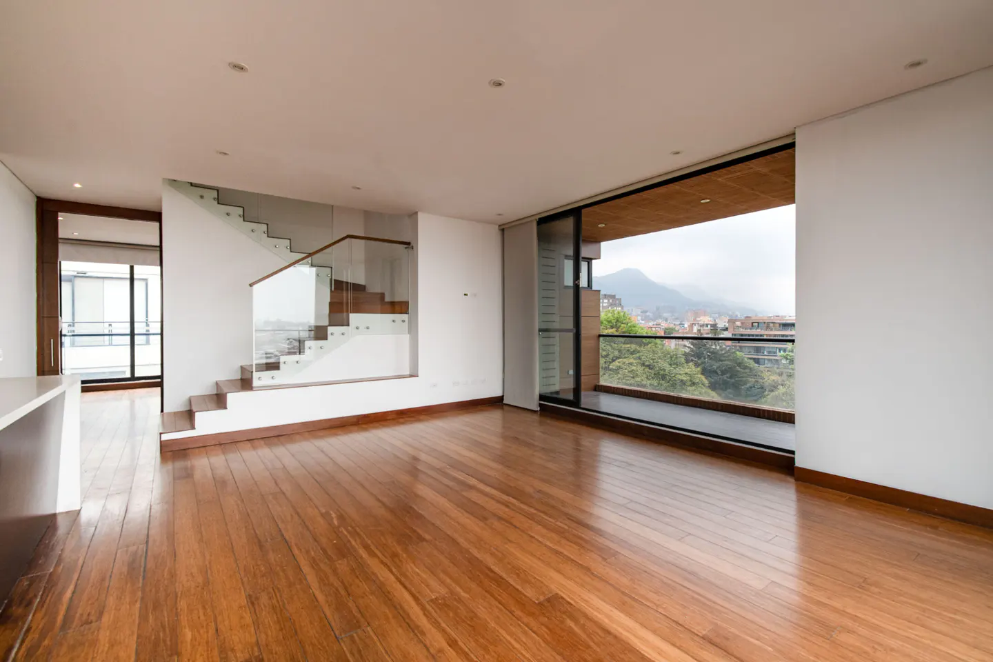 Bright, empty living room with hardwood floors, white walls, and a staircase with glass railings. A large window offers a city view.