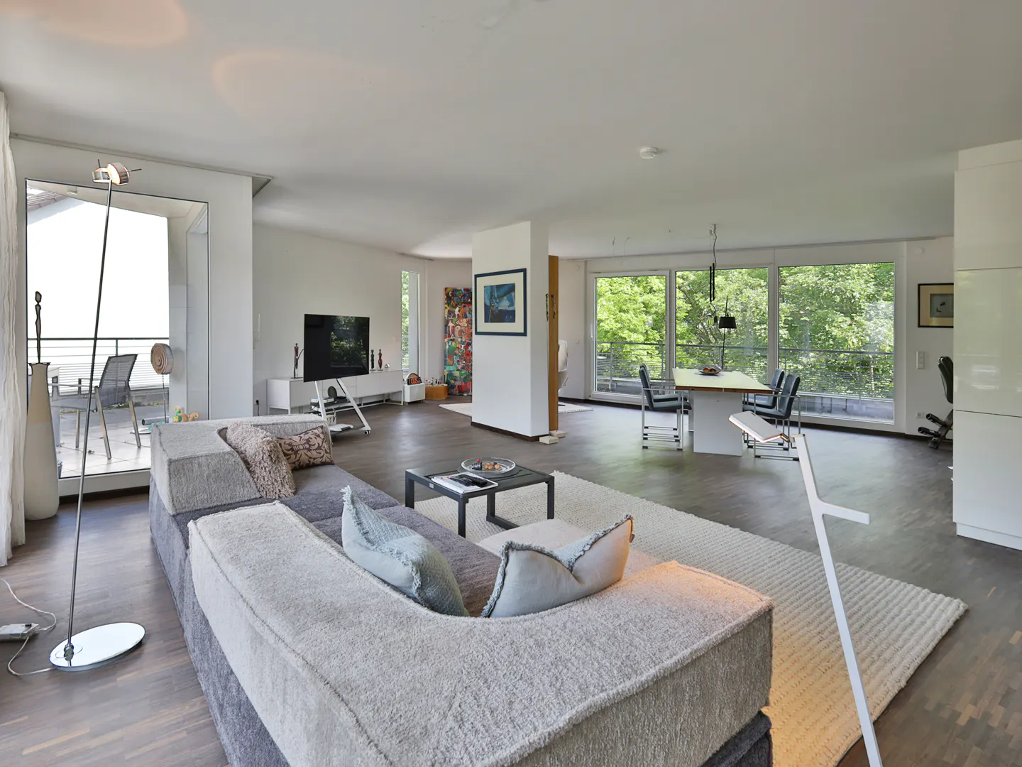 Bright, open-concept living room with a large gray sectional sofa, a black coffee table, and a white rug. Dining table and chairs are visible in the background.